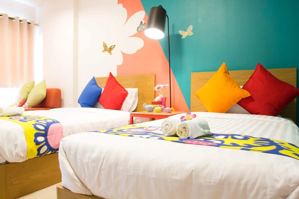 A hotel room with two beds, colorful pillows, a small table with a lamp, and a wall with butterfly decals.
