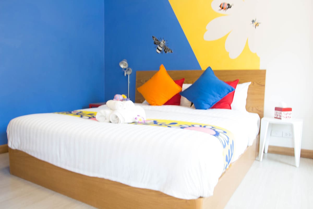 A neatly made bed with colorful pillows, a bedside table with a box, and a wall with a bee and butterfly decal.