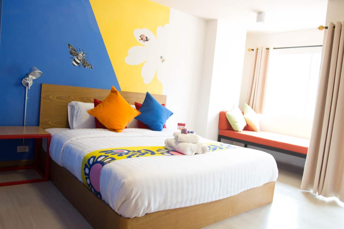 A hotel room with a bed, a nightstand, a lamp, a window with curtains, and a wall with a flower and bee decoration.