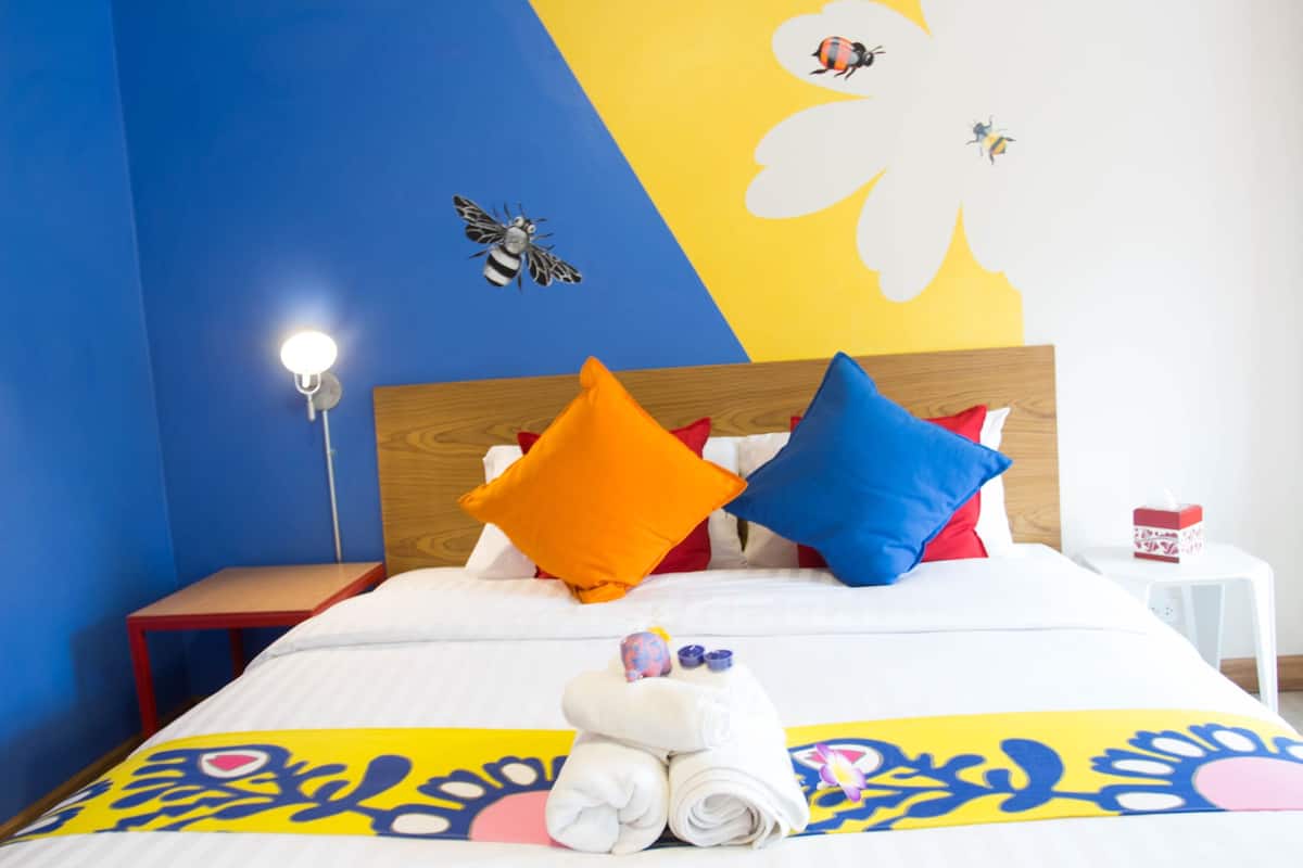 A neatly made bed with colorful pillows, a wall with a bee and flower mural, and a bedside table with a lamp and a tissue box.