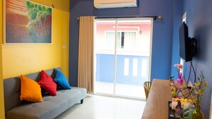 A living room with a sofa, colorful cushions, a painting, an air conditioner, a sliding glass door, and a wooden table with a vase. - Non Nid Non Noi Design Residence (Chiang Mai)
