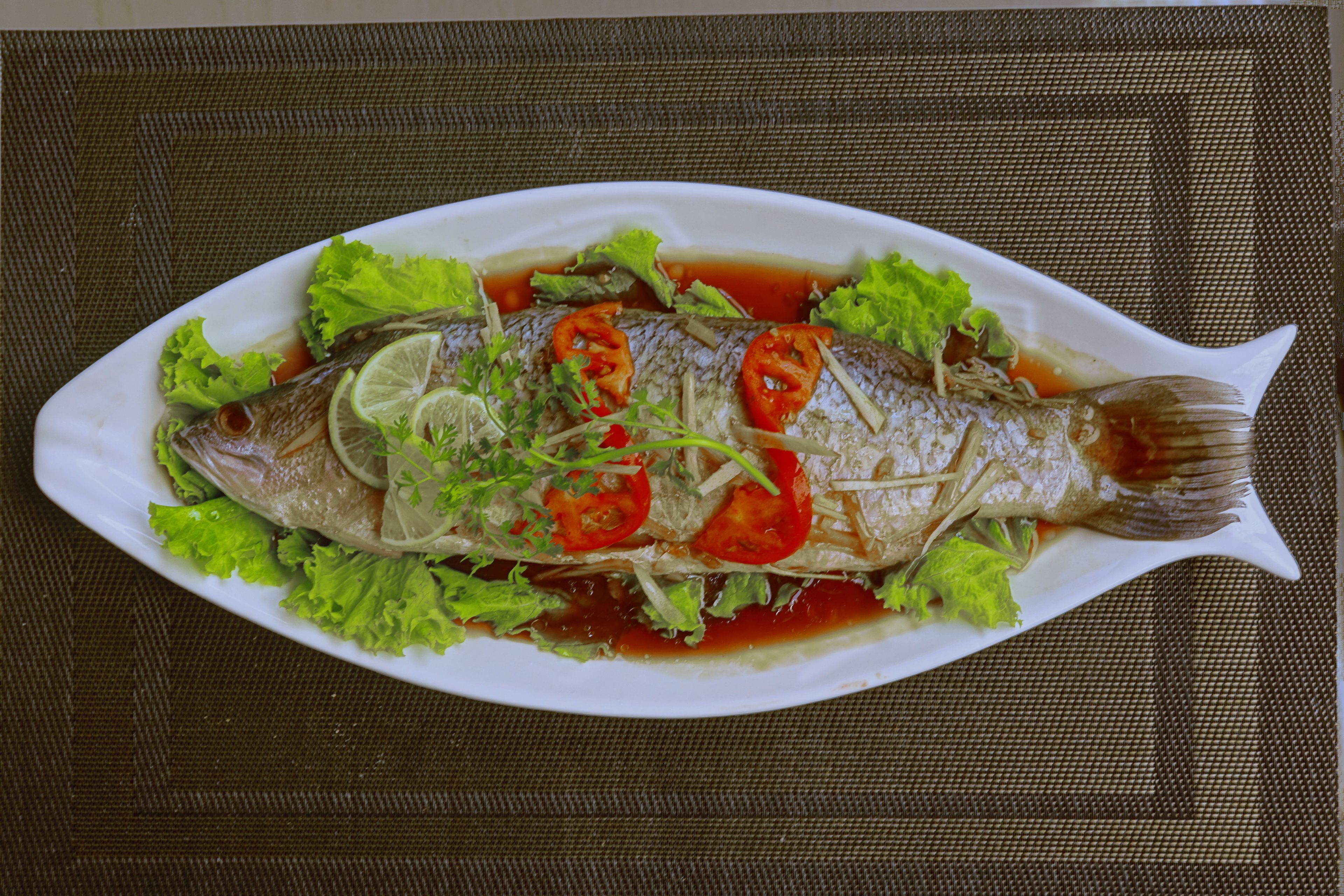 Une assiette ovale blanche sur laquelle est présenté un poisson entier, agrémenté de laitue, de citron vert et de tranches de tomates, le tout servi sur un set de table marron texturé.