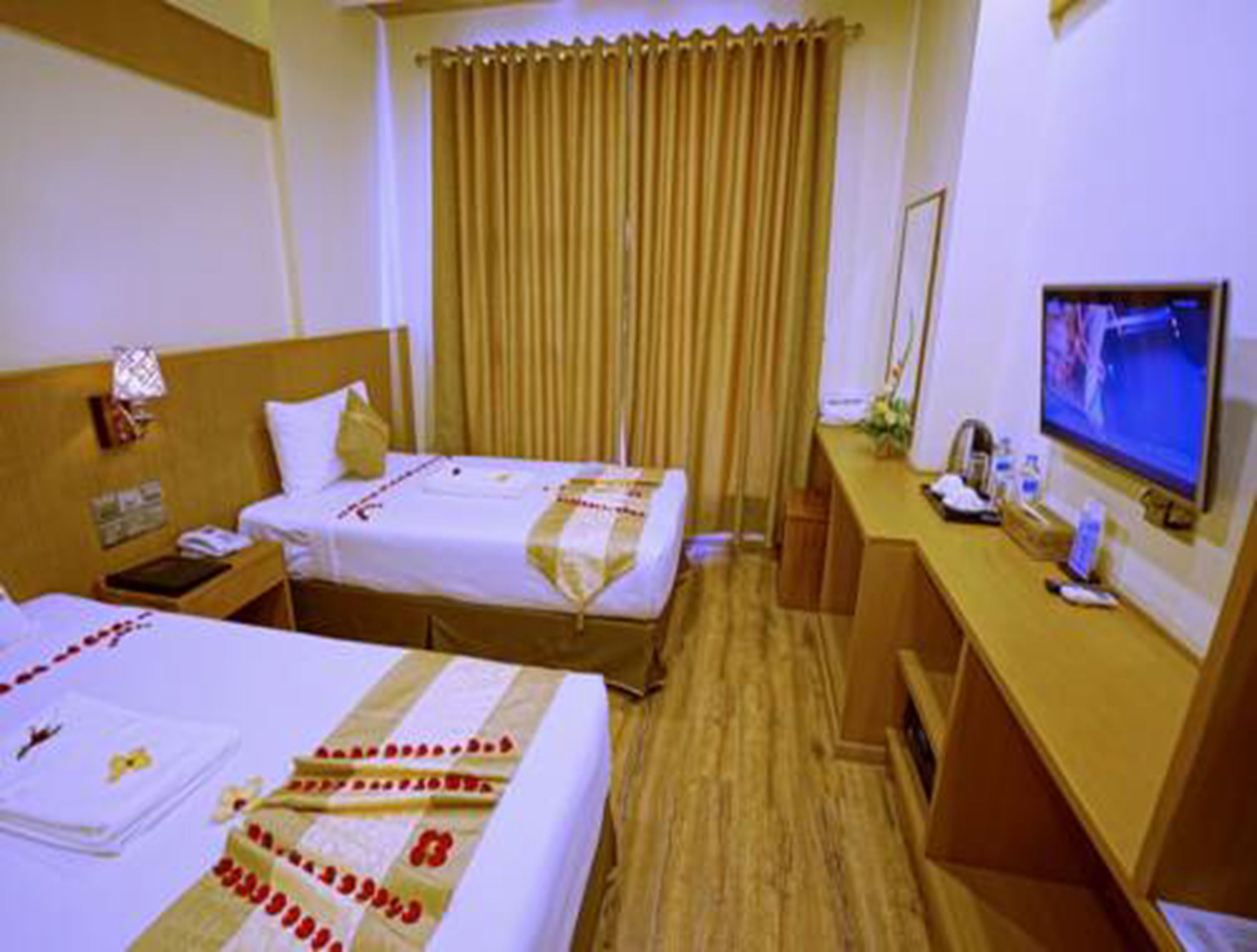 A hotel room with two beds, a desk with a computer and phone, a TV, and a window with curtains.