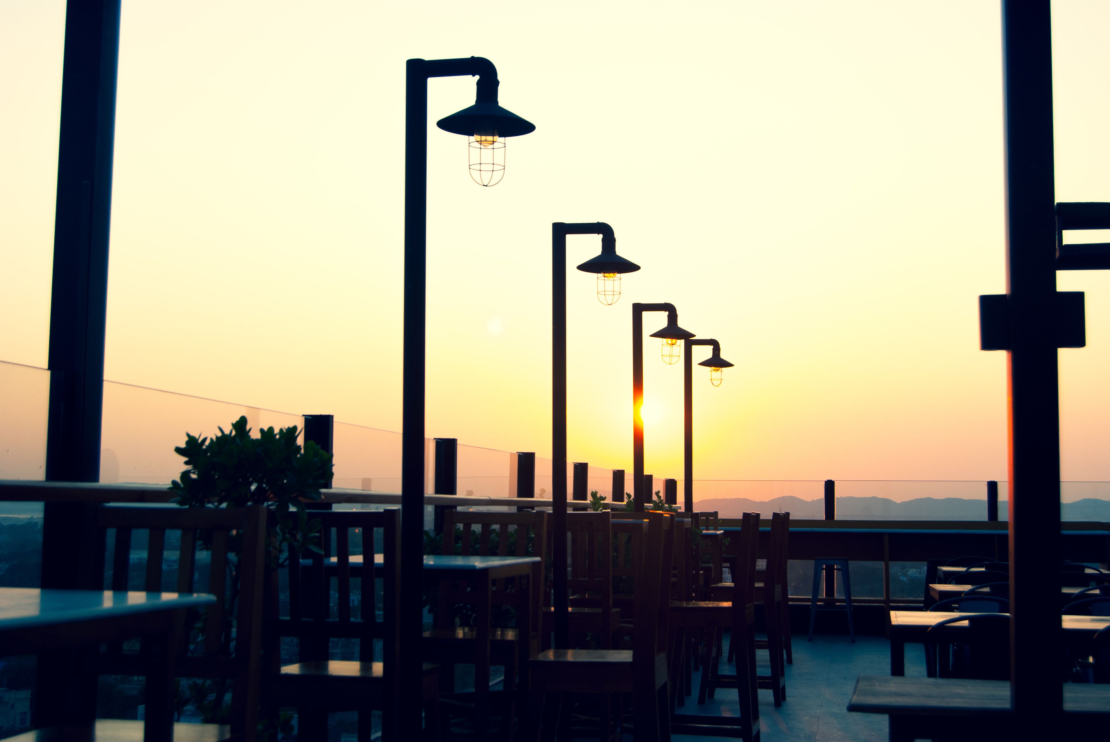 An outdoor seating area with tables and chairs, a row of street lamps, and a sunset view.
