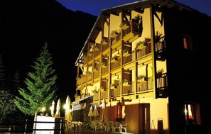 Un hotel de varios pisos con balcones, mobiliario exterior y montañas al fondo.