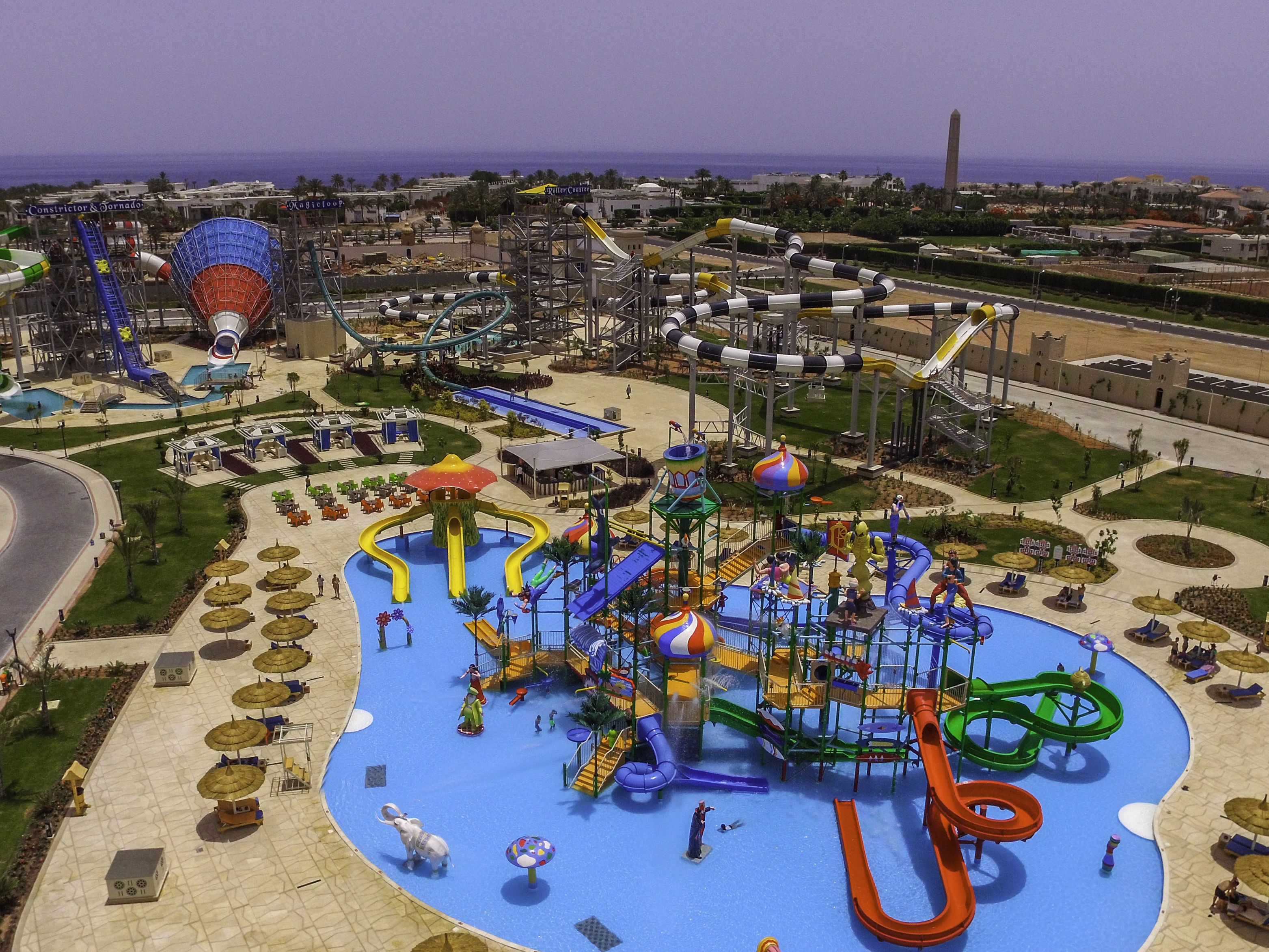 Ein Wasserpark mit zahlreichen Wasserrutschen und Spielbereichen, umgeben von einer begrünten Anlage mit Sitzgelegenheiten und Sonnenschirmen.