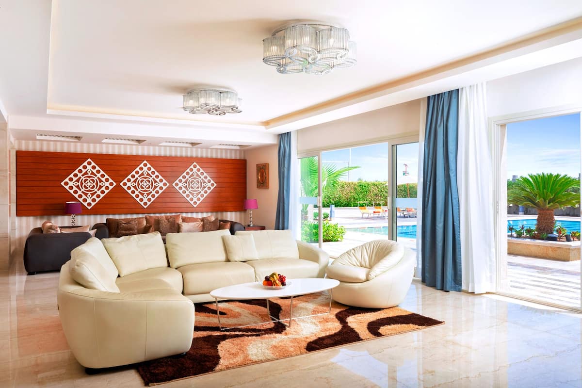 Ein geräumiges Wohnzimmer mit einem beigefarbenen Sofagarnitur, einem Couchtisch und einem gemusterten Teppich. Ein großes Fenster bietet Blick auf einen Pool und Grünflächen.