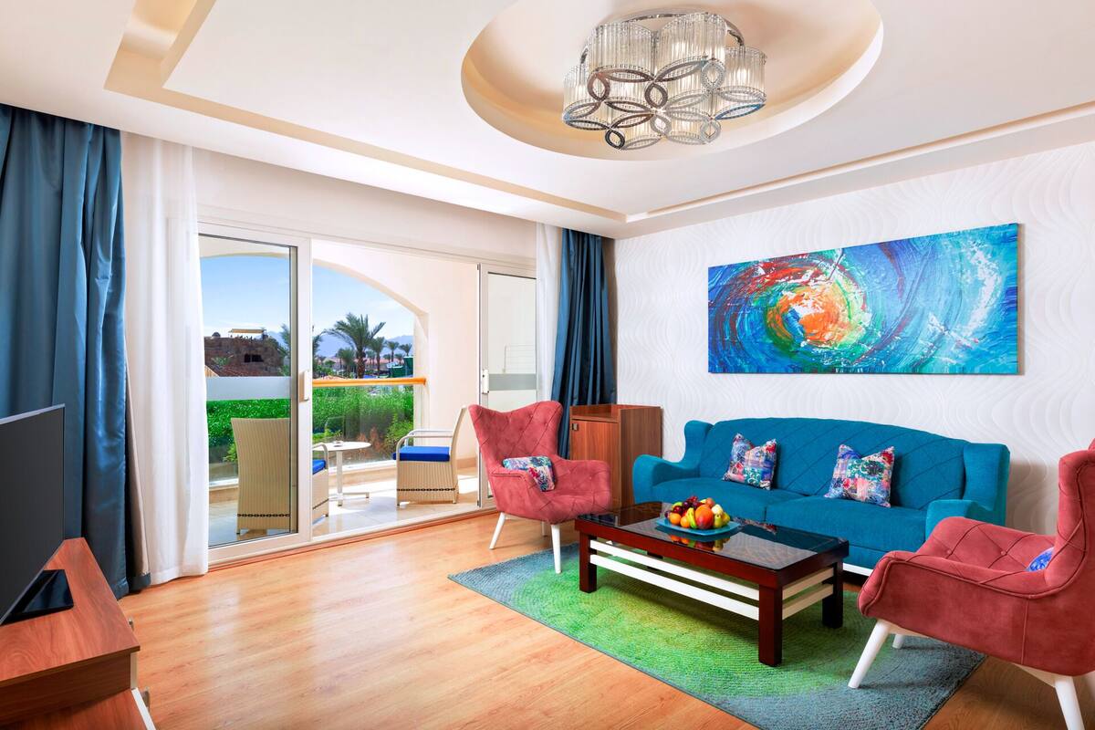 Ein modernes Wohnzimmer mit einem blauen Sofa, roten Sesseln und einem Couchtisch mit Obst. Ein großes abstraktes Gemälde hängt an der Wand, und es gibt einen Blick auf einen Balkon mit viel Grün.