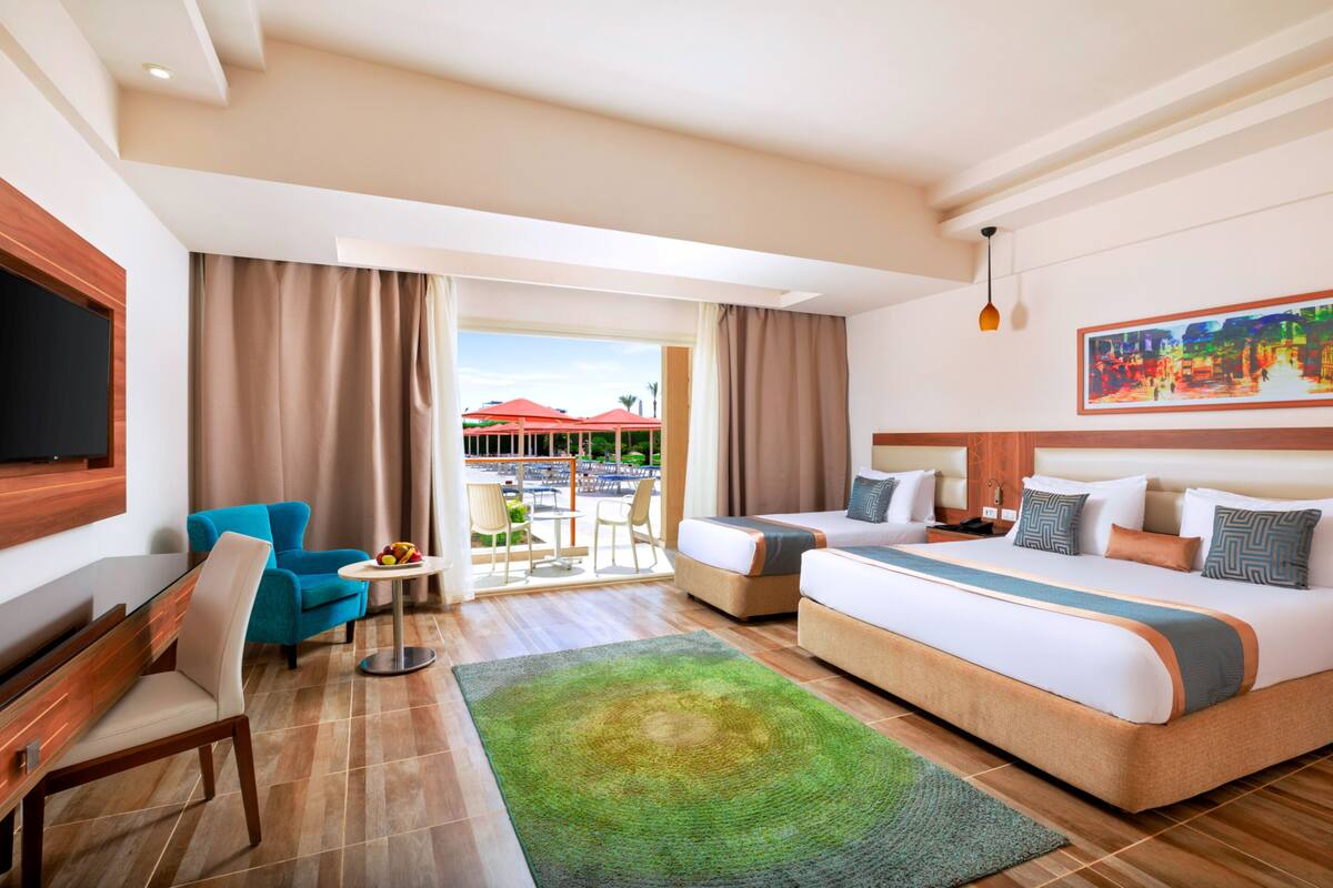 Ein Hotelzimmer mit einem großen Bett, einem Schreibtisch mit Stuhl, einem kleinen Tisch mit Stühlen und einem Balkon mit Blick auf Gebäude und Palmen.