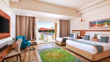 Habitación de hotel con una cama grande, un escritorio con una silla, una pequeña mesa con sillas y un balcón con vistas a edificios y palmeras.