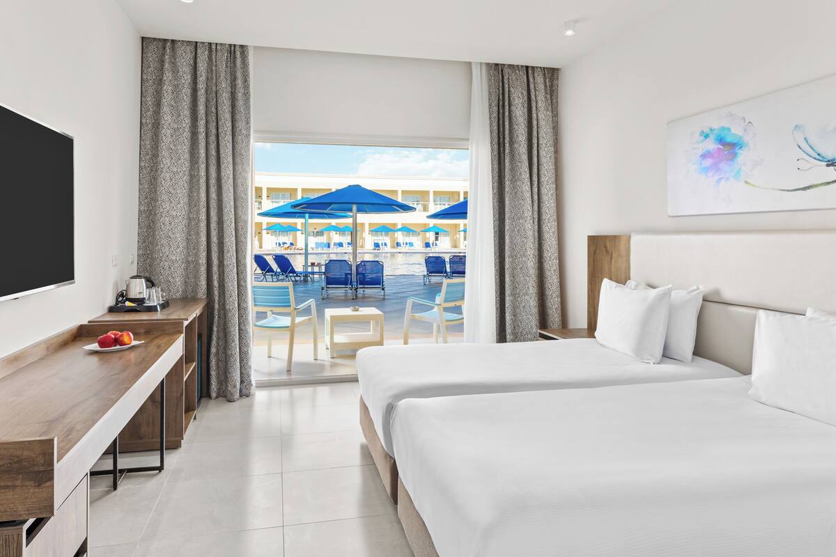 Ein Hotelzimmer mit Bett, Schreibtisch und Blick auf einen Poolbereich mit Sonnenschirmen.
