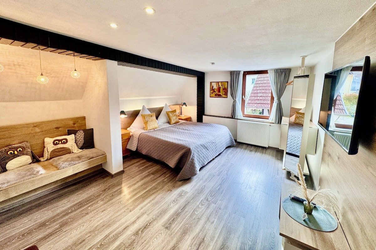Un dormitorio moderno con una cama grande, mesitas de noche, un escritorio con computadora y una ventana con cortinas.