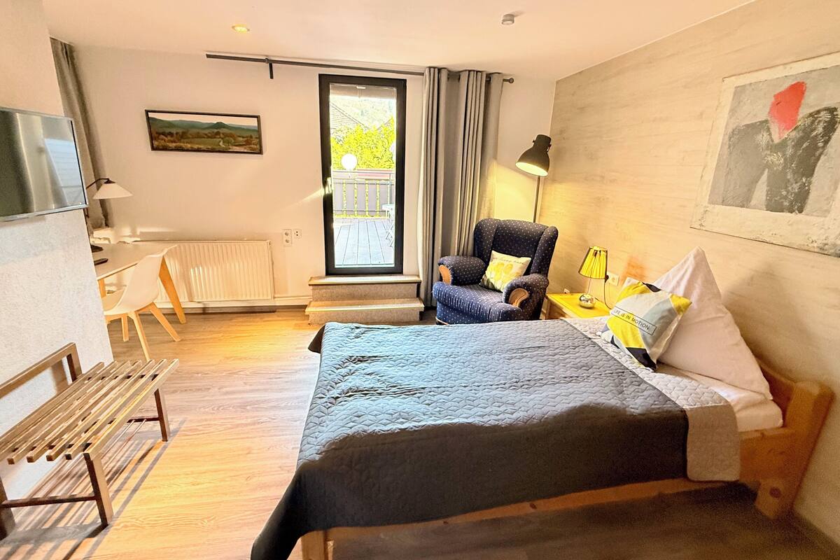 Una habitación de hotel con cama, silla, escritorio, televisor y ventana con cortinas.