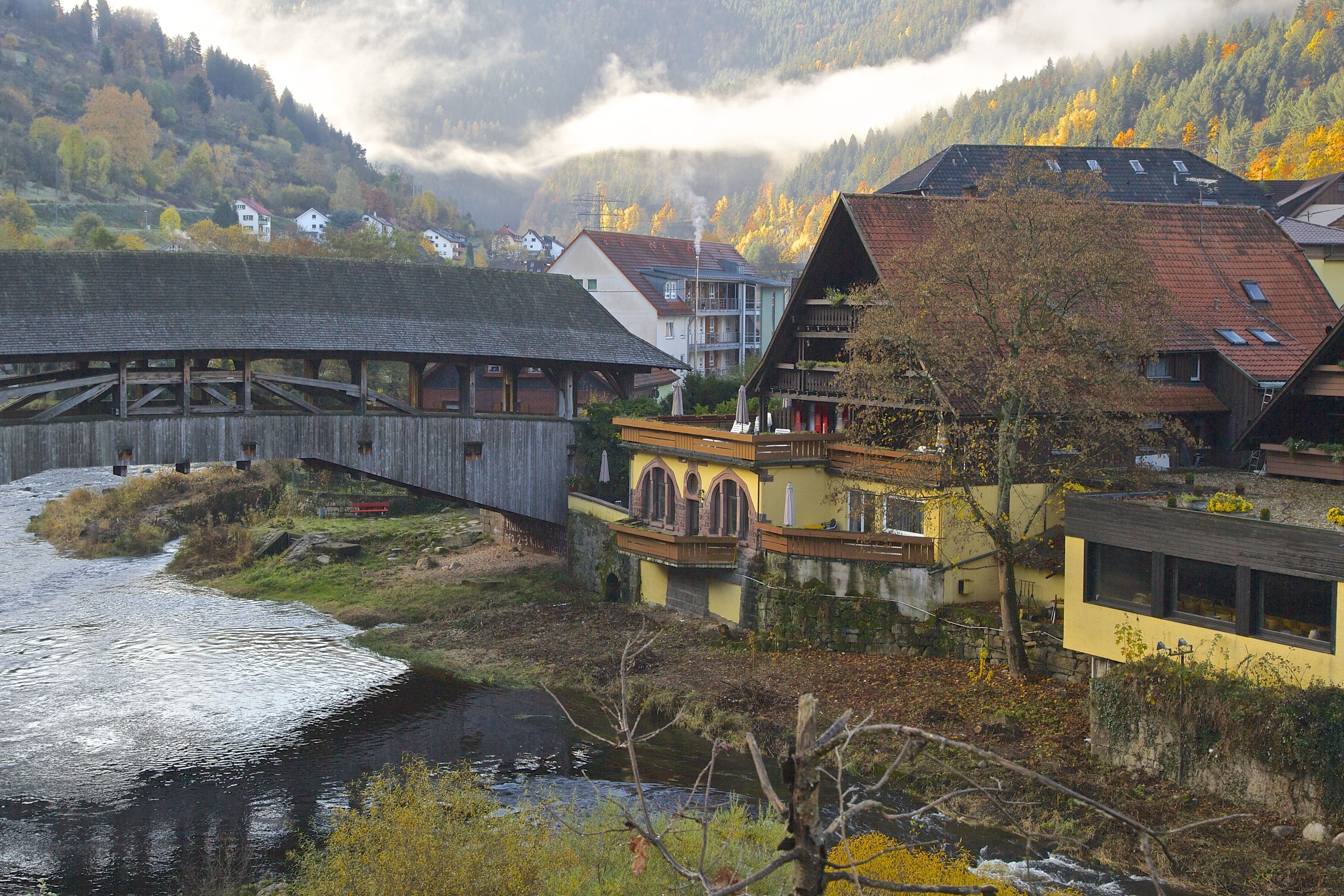 A wooden bridge over a river, surrounded by houses with balconies and a mountain backdrop.