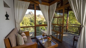 A room with a wooden sofa, a table with a fruit bowl, and a view of a mountainous landscape through large windows. - Popa Garden Resort (Kyaukpadaung)