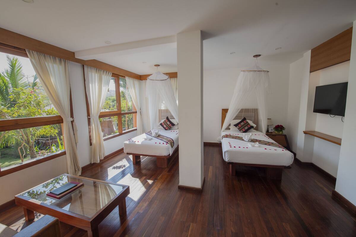 Habitación de hotel con dos camas, televisión, mesa y ventanas amplias con cortinas.