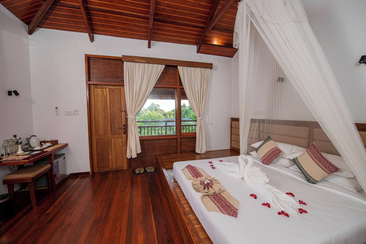 Un dormitorio con piso de madera, una cama con ropa blanca y almohadones decorativos, un escritorio de madera con una silla y una ventana con cortinas.