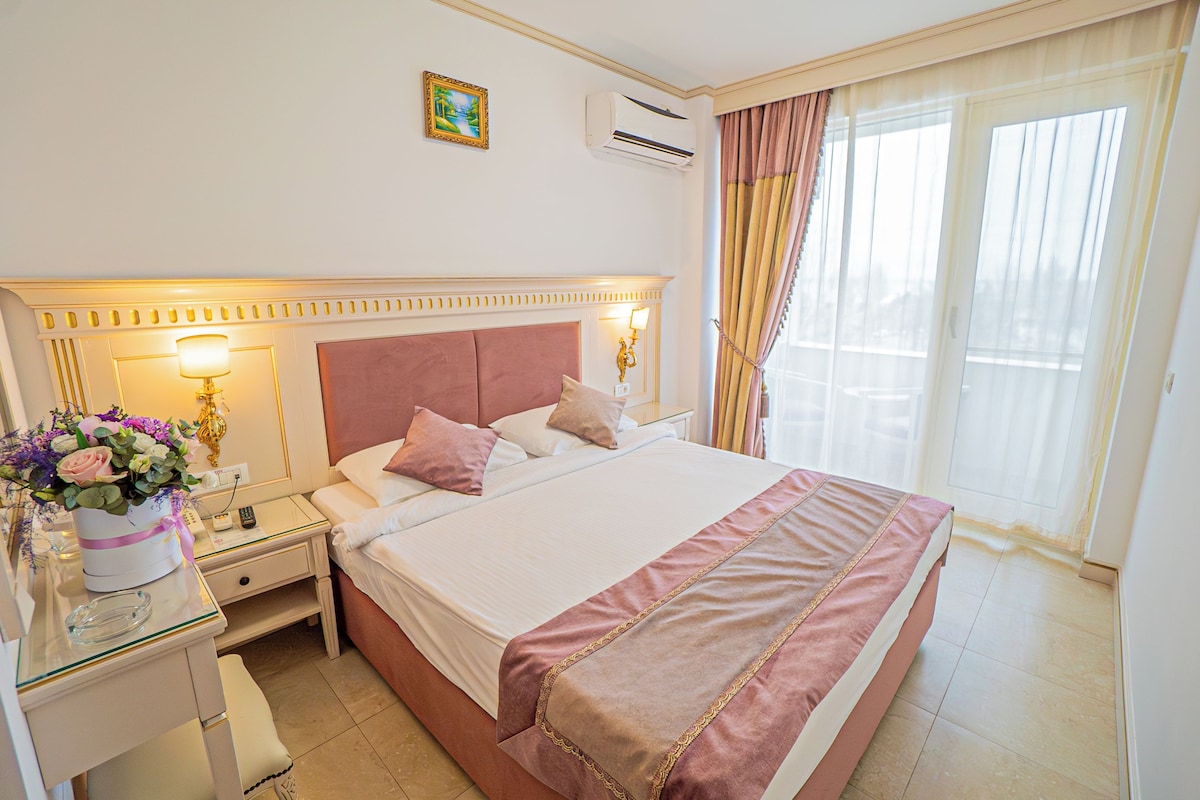 A hotel room with a large bed, bedside tables, a nightstand, a lamp, a vase with flowers, and a window with curtains.