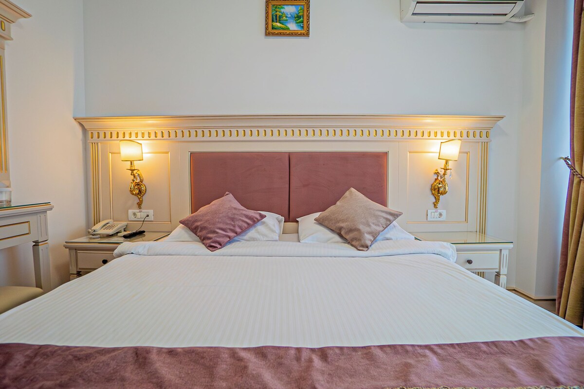 A hotel room with a large bed, two bedside lamps, a telephone, and a painting on the wall.
