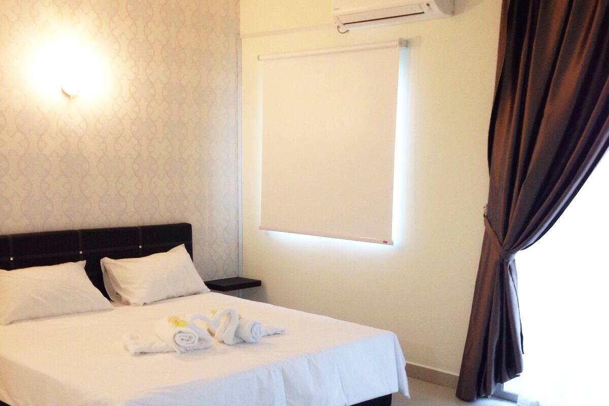A hotel room with a large bed, a bedside table, an air conditioning unit, a window with curtains, and a wall-mounted light.