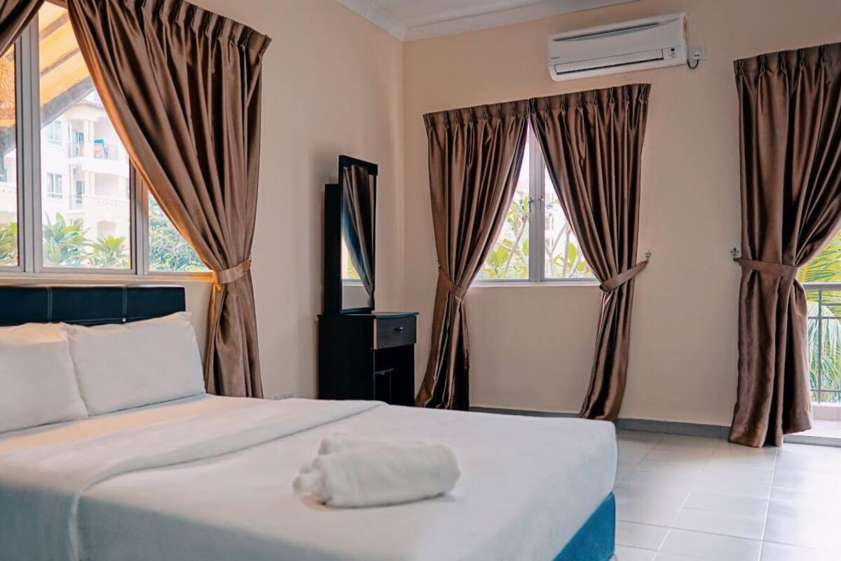 A hotel room with a bed, curtains, an air conditioner, and a view of buildings outside.