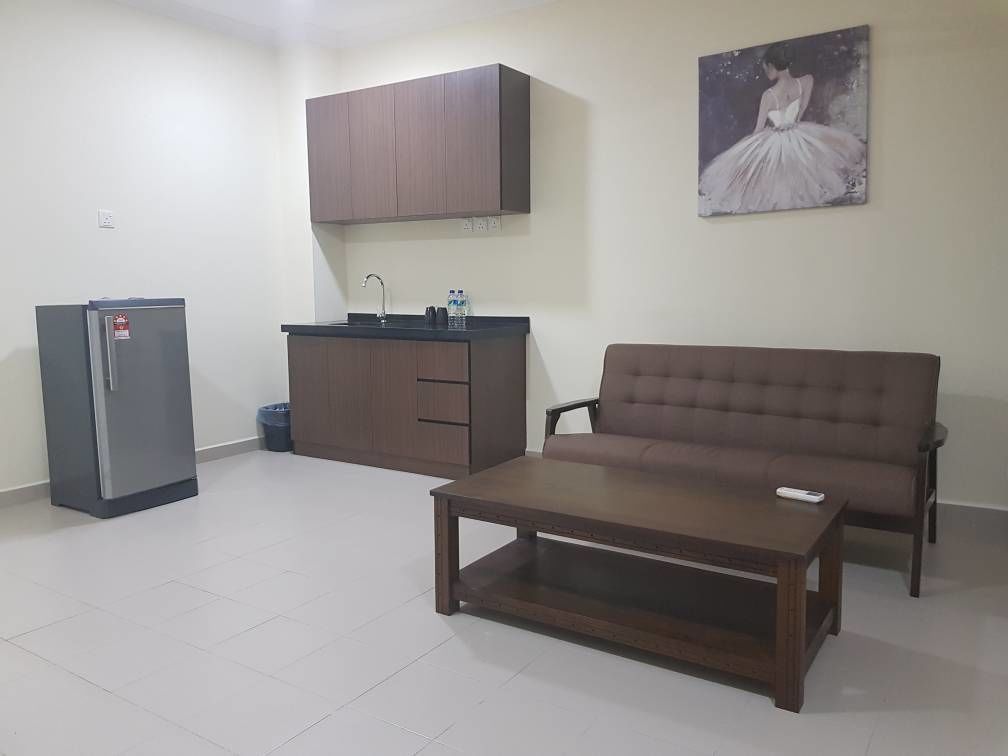 A living room with a brown sofa, a wooden coffee table, a grey refrigerator, and a kitchen area with cabinets and a sink.