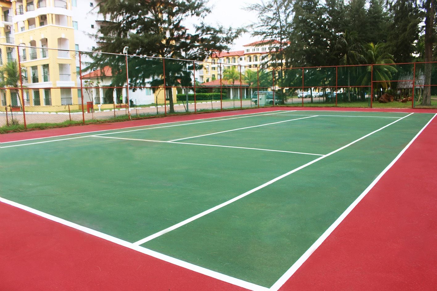 Cancha de tenis exterior con superficie roja y verde, rodeada por una valla y árboles.