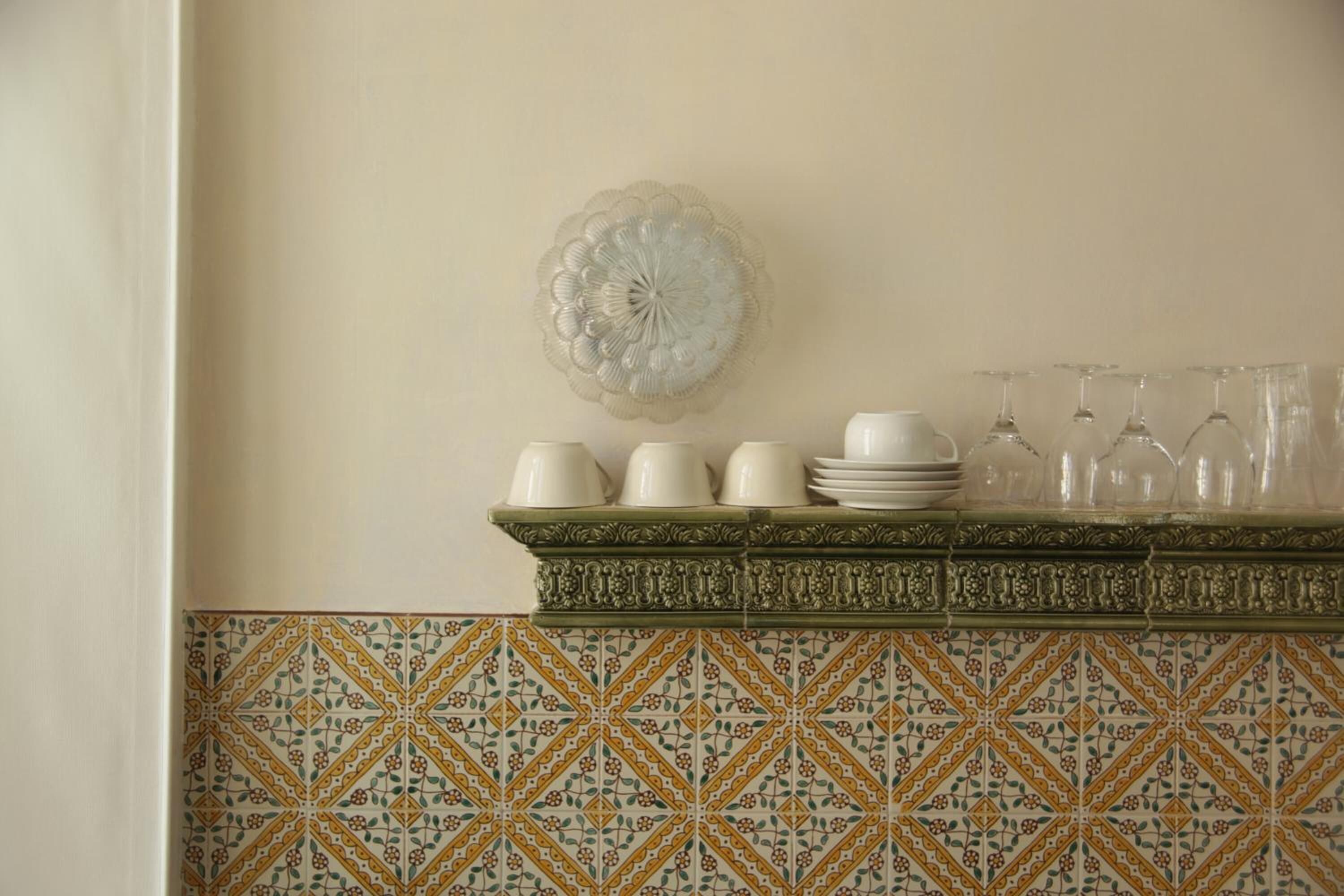 Una cocina con un salpicadero decorativo, un plato de vidrio montado en la pared y un estante con tazas, platos y copas de vino blancos.