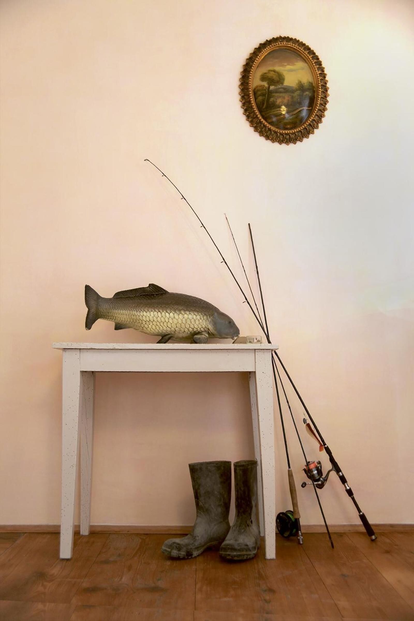 Un pez sobre una mesa, cañas de pescar y botas junto a una pared.