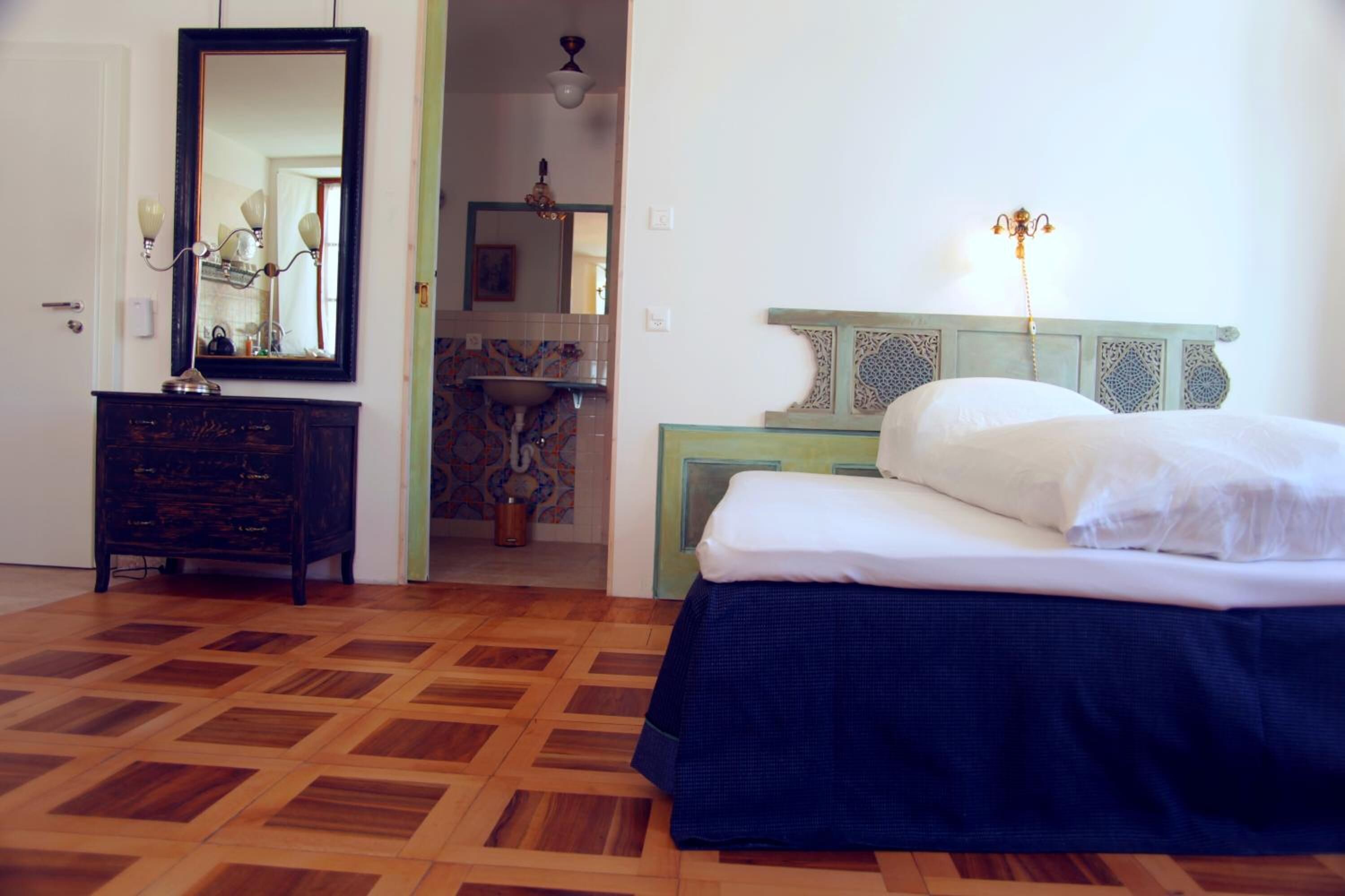 Un dormitorio con piso de parquet de madera, una cama con cubrecama azul, un aparador con espejo y una mesita de noche verde con lámpara.