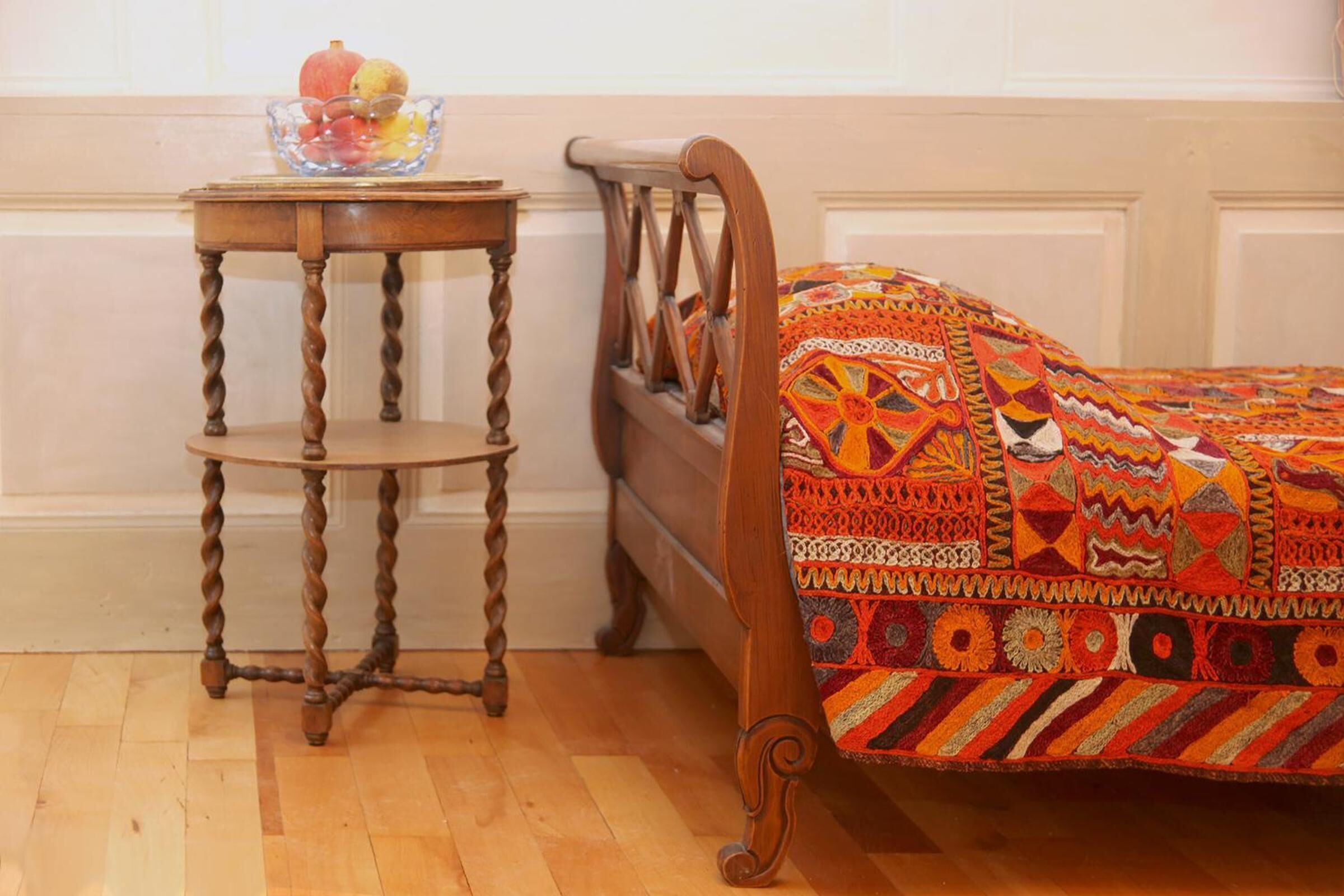 Uma cama de madeira com um edredom colorido estampado, uma mesinha de cabeceira de madeira com uma tigela de frutas e piso de madeira.