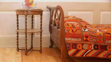 Uma cama de madeira com um edredom colorido estampado, uma mesinha de cabeceira de madeira com uma tigela de frutas e piso de madeira.