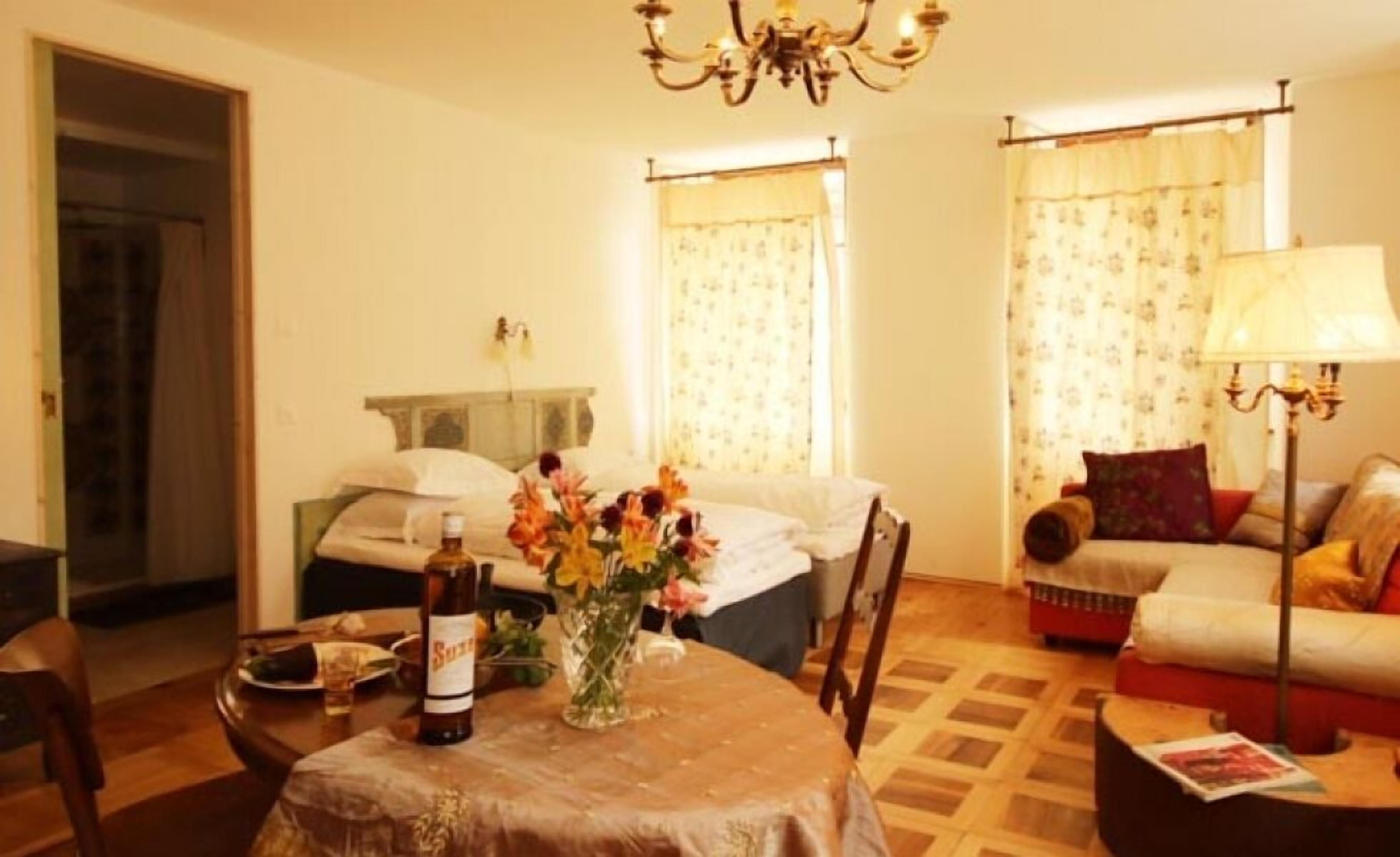 A hotel room with a bed, a dining table with a bottle, a vase with flowers, a sofa, and a lamp.