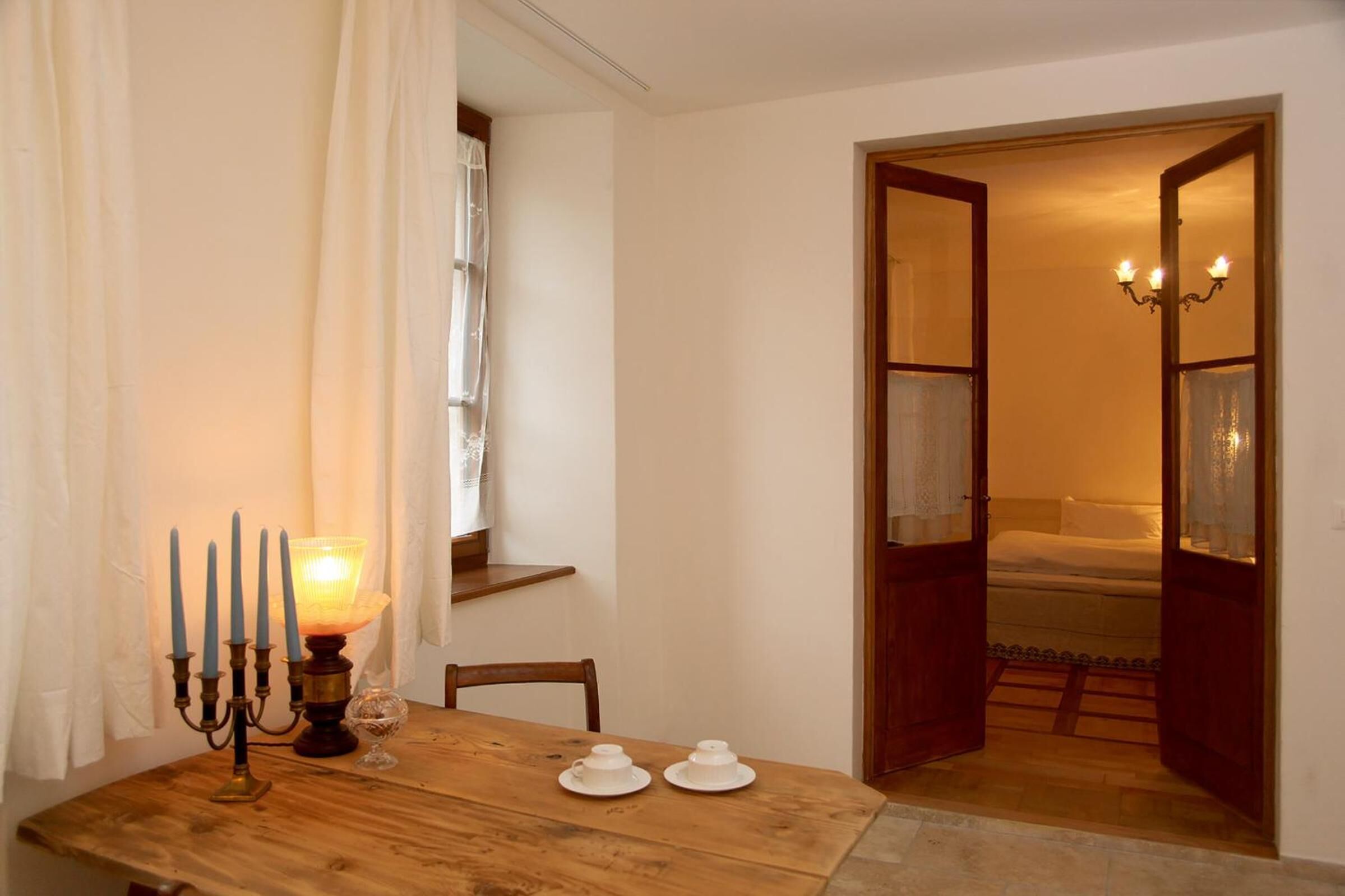 A wooden dining table with a candelabra and two teacups, a wooden chair, and a staircase leading to a bedroom with a bed and a chandelier.