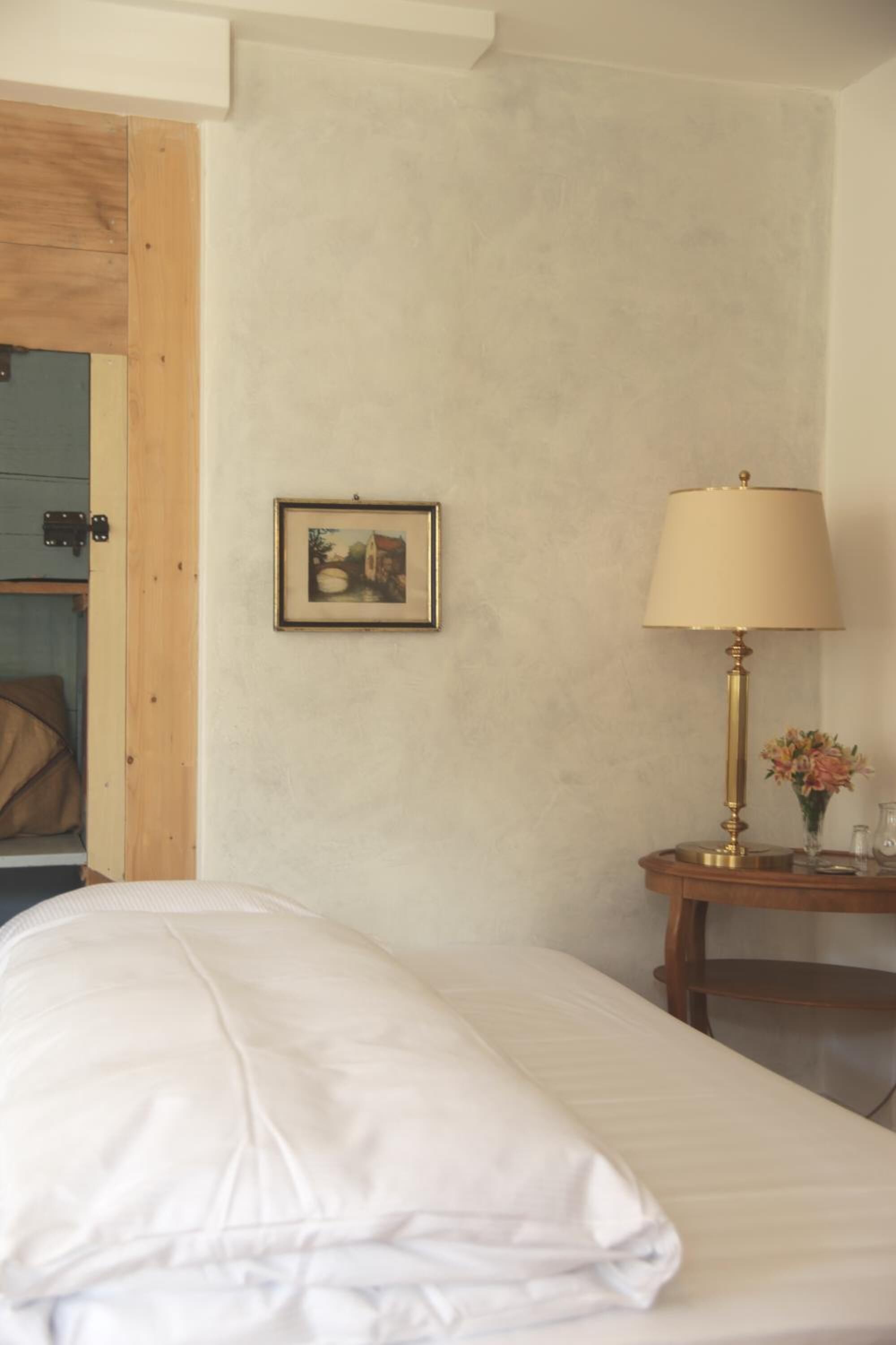 Una cama bien tendida con edredón blanco, un cuadro en la pared, una lámpara en una mesita de noche y un marco de puerta de madera.