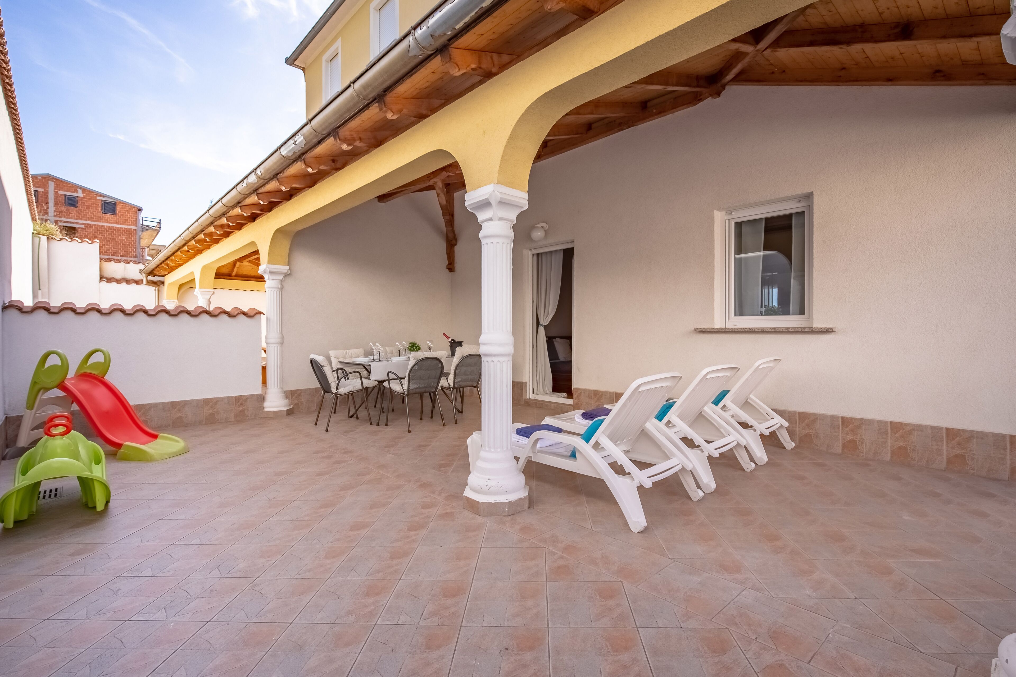 An outdoor patio with a tiled floor, a children's slide, lounge chairs, and a table with chairs.