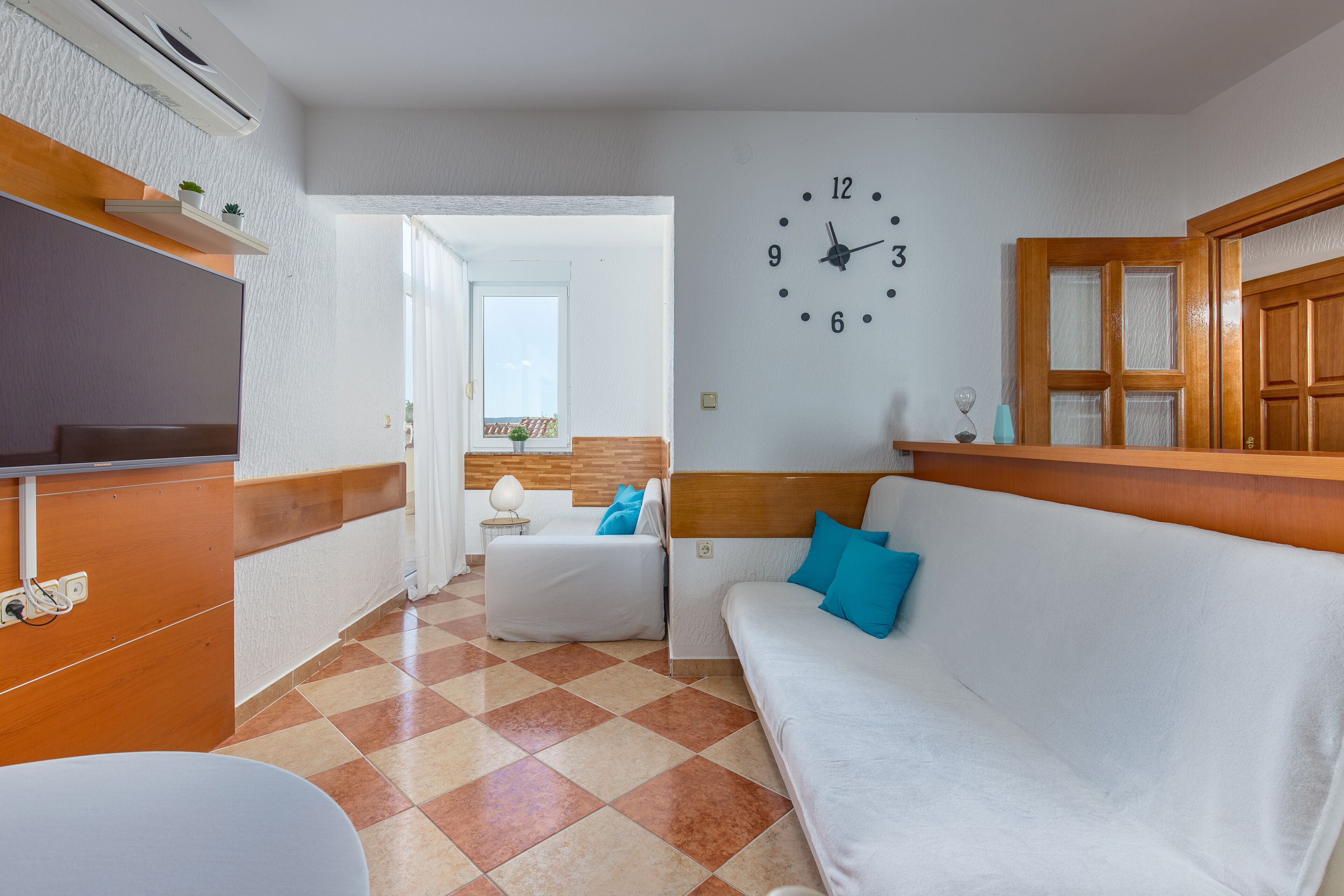A hotel room with a checkered floor, a white sofa with blue pillows, a wooden headboard, a wall clock, and a television on a wooden cabinet.