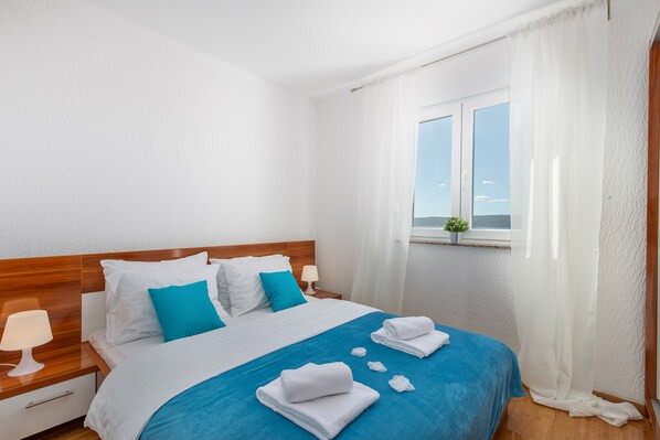 Habitación de hotel con cabecera de madera, ropa de cama blanca y azul, una ventana con vistas y una pequeña planta en el alféizar.