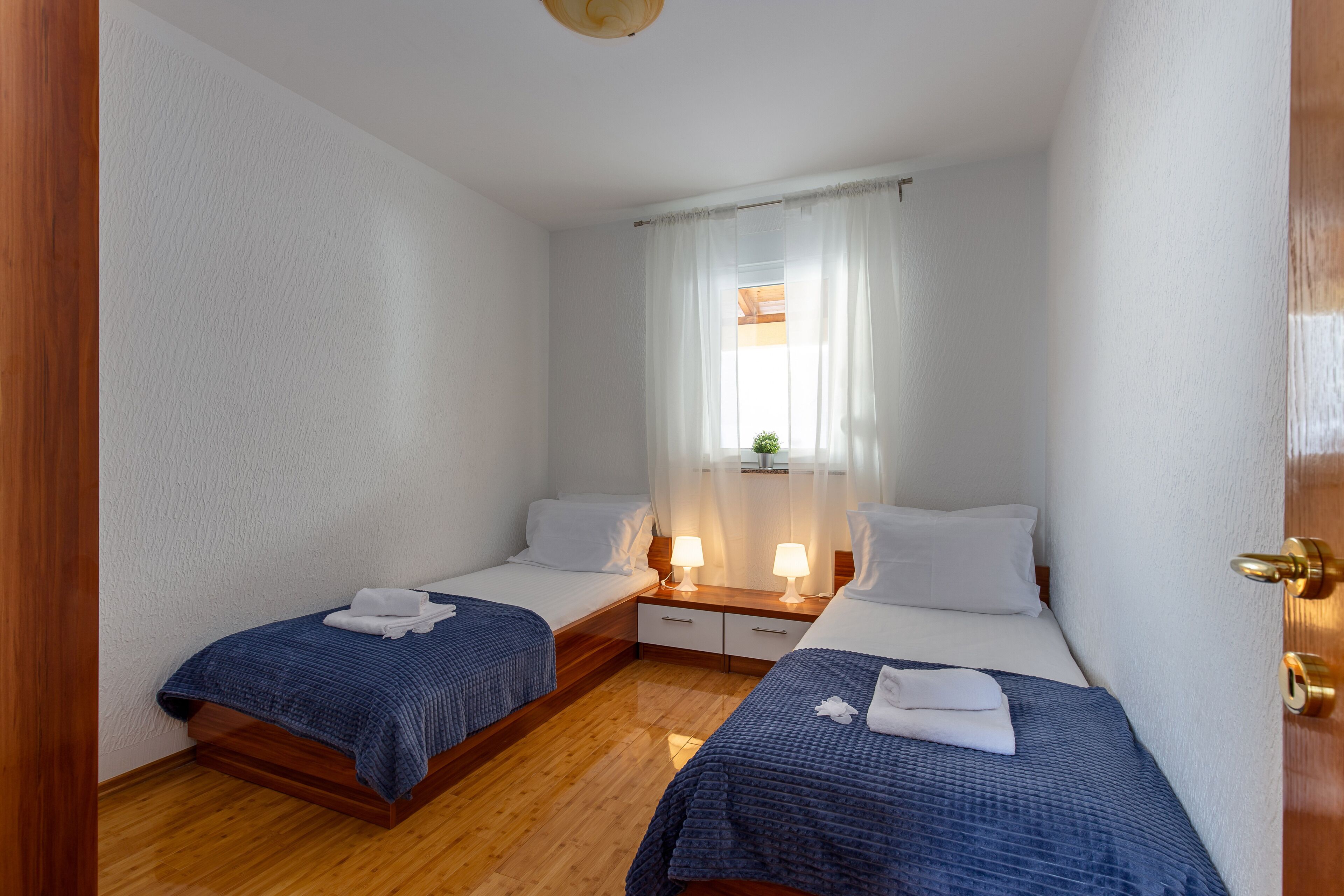 A room with two beds, wooden flooring, white walls, and a window with sheer curtains.