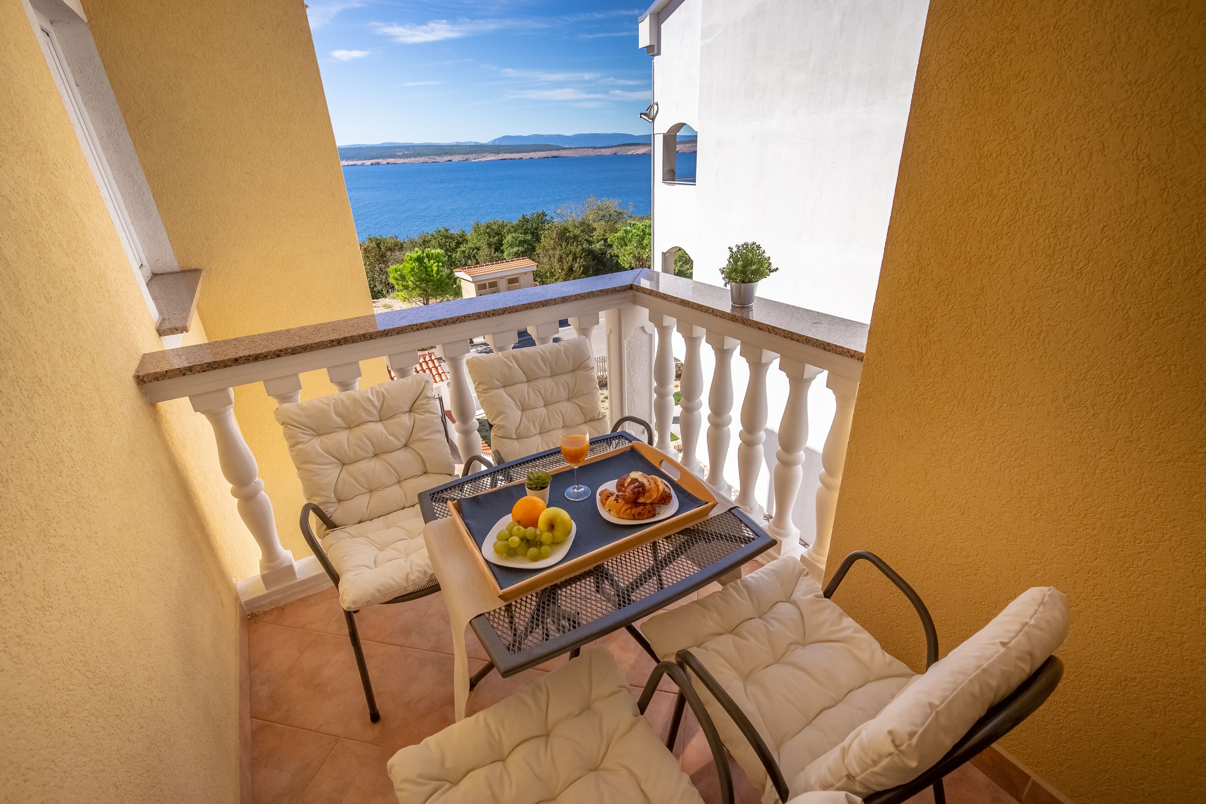 A balcony with a table set for breakfast, including grapes, an orange, and croissants, overlooking a coastal view.