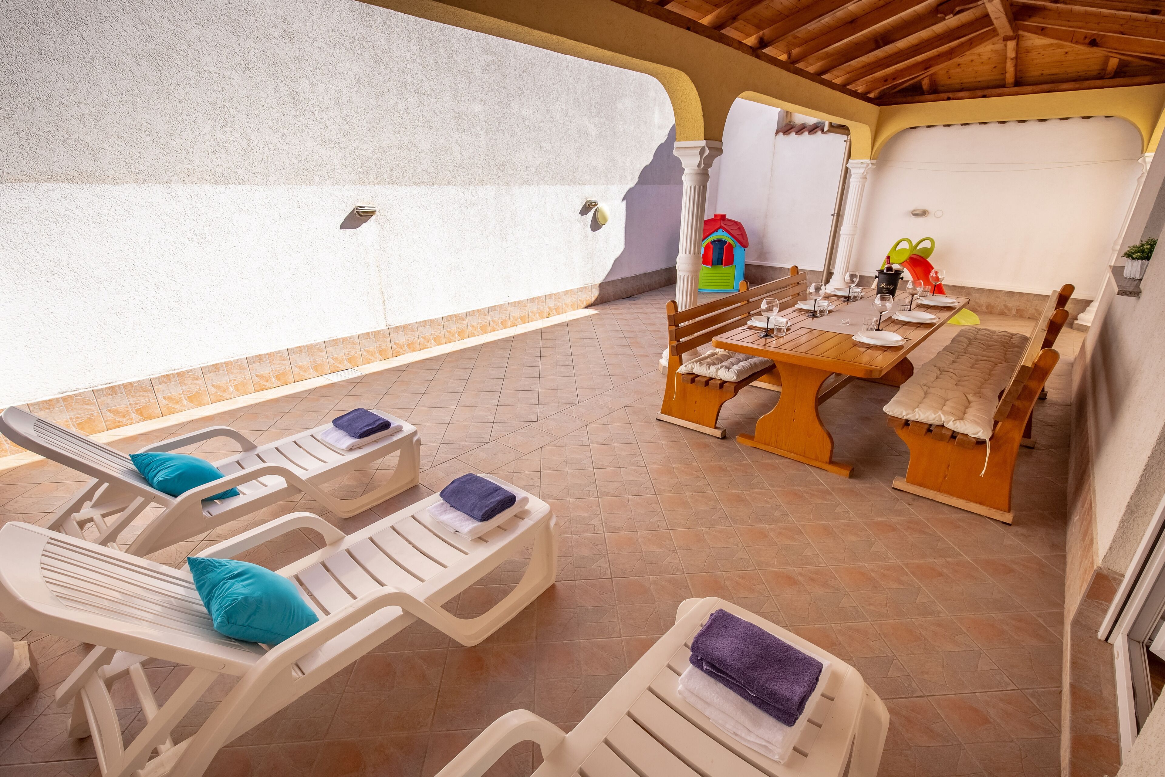 A sunlit patio with lounge chairs, a dining table set for four, and a colorful children's playhouse.