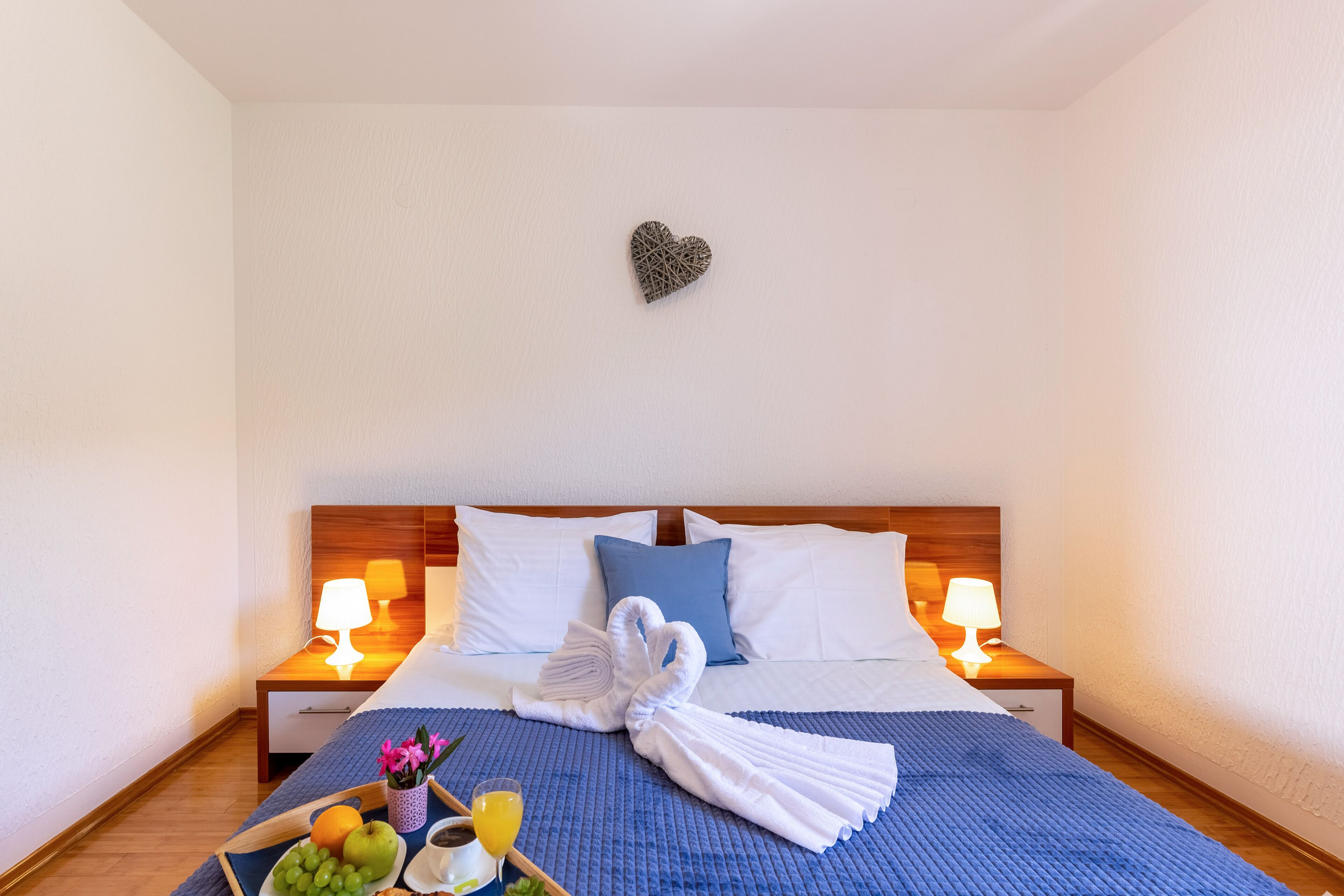 Una cama bien hecha con una colcha azul, dos lámparas de noche, una decoración con forma de corazón en la pared, y una bandeja con frutas y un vaso de jugo en la mesita de noche.