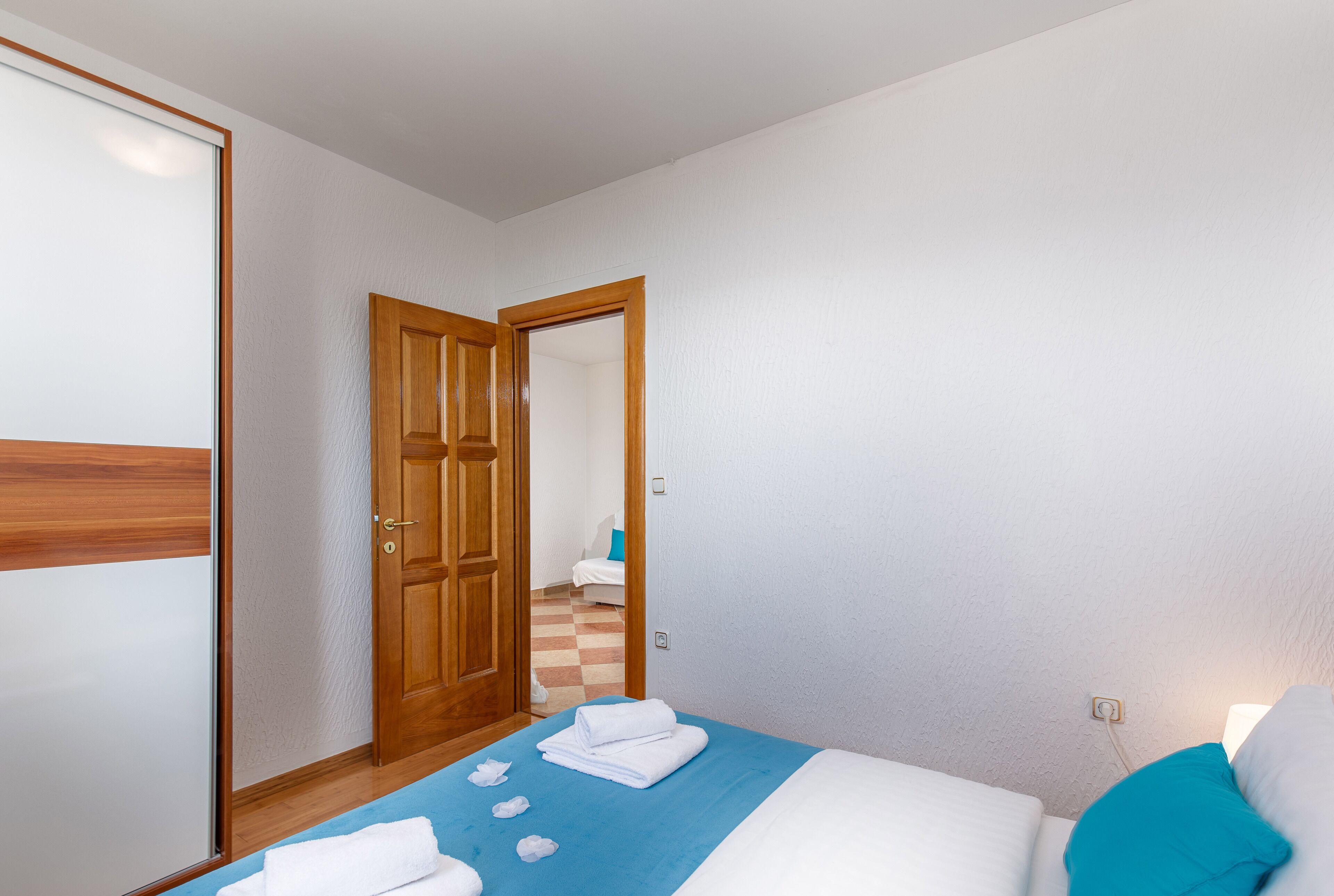 A bedroom with a bed, a blue bedspread, white towels, a wooden door, and a view into another room.
