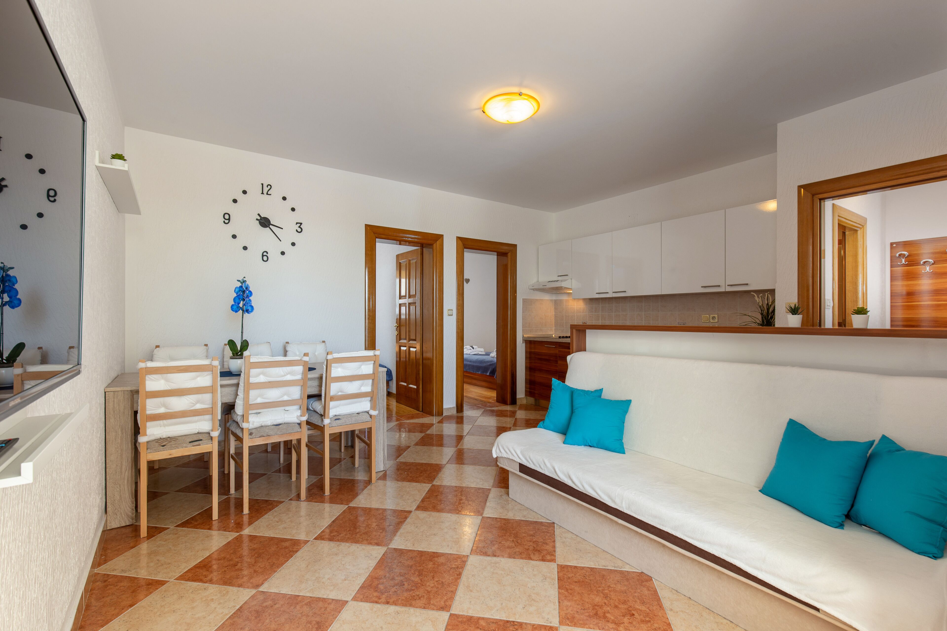 A modern living room with a checkered floor, a white sofa with blue cushions, a dining area with wooden chairs, and a kitchen in the background.
