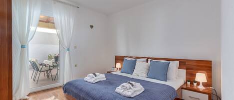 A bedroom with a wooden headboard, a bed with blue bedding, two bedside tables with lamps, and a view of an outdoor seating area through a glass door.