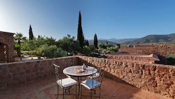Um pátio com mesa e cadeiras redondas, rodeado por paredes de pedra e vegetação, com vista para uma paisagem montanhosa.