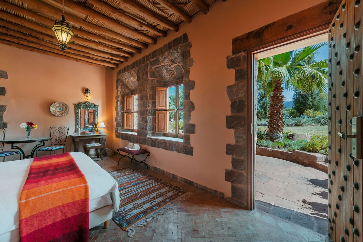 Habitación con cama, una mesa, una silla, ventana y vista a palmeras, además de una pared de piedra.