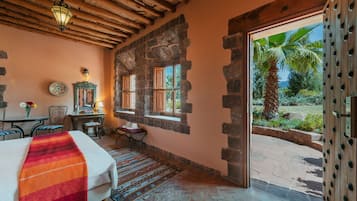 Habitación con cama, una mesa, una silla, ventana y vista a palmeras, además de una pared de piedra.