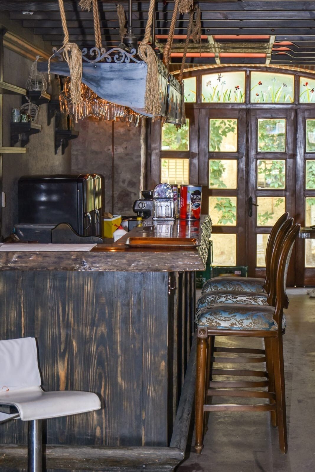 Una zona de bar rústica con mostrador de madera, sillas de estilo vintage y un amplio ventanal con vistas a la vegetación.