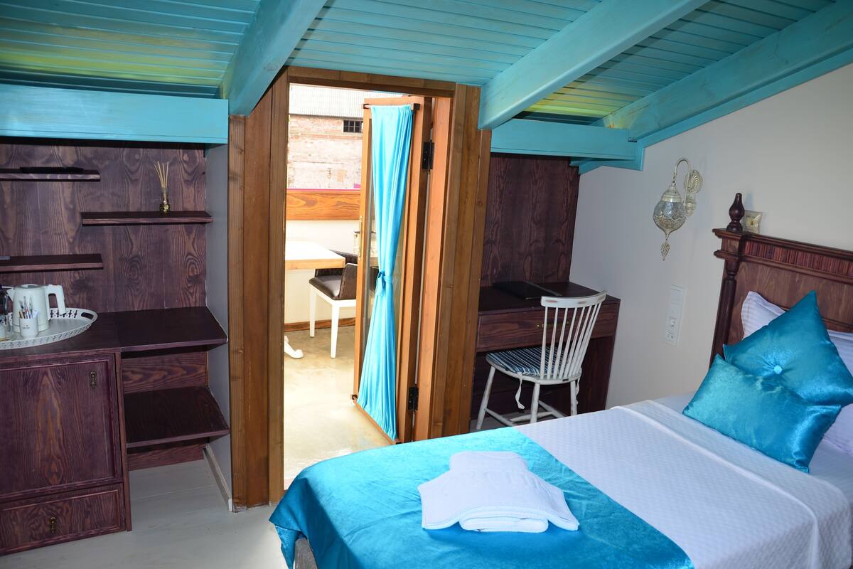 A room with a bed, a desk, and a chair. There is a window with a curtain, a wall-mounted shelf, and a wall clock.